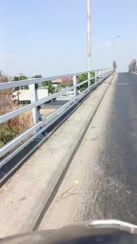 The video is a first-person perspective shot, seemingly from a bicycle or motorcycle, ascending a concrete bridge. The railing on the left side of the bridge is made of white metal bars. Below the bridge, there is a road with some vehicles, including a white truck and a black car. The environment appears to be an urban or suburban area with trees and buildings visible in the distance. The sky is clear and blue. The bridge surface is made of asphalt and appears worn. There are a few rocks or debris on the edge of the bridge. The camera is moving forward and upward.