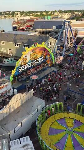 This aerial drone footage captures a bustling amusement park scene in Gloucester, United States of America. The primary subjects are the various carnival rides and the throngs of people enjoying them. We see a large pendulum ride swinging back and forth, a Ferris wheel-like ride with cars suspended on a rotating circular track, and a large, colorful ride with a central spinning platform. The "Seven Seas" ride, a prominent attraction with a large, illuminated sign depicting a pirate ship on a wave, is also visible.

The event taking place is clearly a carnival or amusement park day. People are seen walking around, queuing for rides, and enjoying the atmosphere. The time of day appears to be late afternoon or early evening, with the golden light of the setting sun casting long shadows and bathing the scene in a warm glow. The weather seems pleasant, with clear skies and a few wispy clouds.

Notable details include the American flags flying from the top of one of the rides, adding a patriotic element. The background reveals glimpses of a coastal town with buildings, piers, and boats, reinforcing the Gloucester location. The overall mood conveyed is one of excitement and fun, typical of a lively carnival. The visible text includes "SEVEN SEAS" on the marquee of one of the rides.