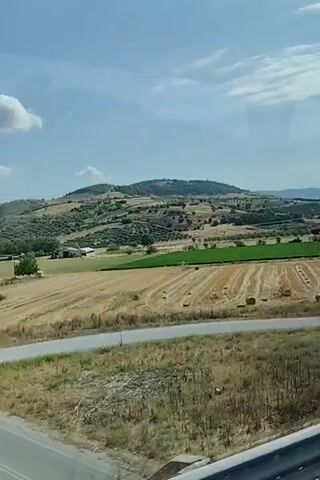 The video shows a view from a moving vehicle of a rural landscape under a blue sky with scattered clouds. The terrain is rolling hills covered in fields, some green and some golden brown, suggesting agriculture. There are scattered trees, including what appear to be olive groves on the hillsides. A road with a fork is visible in the foreground. In the distance, there are some buildings and a tall electrical pylon. The overall impression is of a sunny day in a countryside setting.