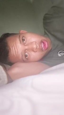 The video shows a close-up, sideways shot of a person's face. The person is lying down on what appears to be a bed with white sheets and a pillow. They are wearing a dark-colored shirt. The lighting is dim, and the background is a dark, solid color. The person's eyes are open, and their mouth is slightly ajar, as if they are speaking or reacting to something. The camera appears to be held by the person themselves, indicated by the first-person perspective and the slight movement of the camera. The video lasts for a few seconds, showing very little change in the person's expression or position.