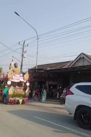 A parade float decorated with red and white fabric and elaborate floral arrangements moves down a street. A large statue of a person in traditional clothing stands on the float. Several people are on the float, some dressed in traditional costumes. A man on a motorcycle drives past the float. In the background, people stand on the side of the street, and buildings line the road. The sky is clear and blue.