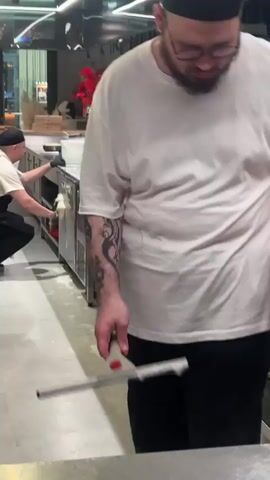 The video shows two people in a kitchen environment. In the foreground, a person wearing a white t-shirt and a black baseball cap is blurred and appears to be holding a cleaning tool or a squeegee, moving it across a surface. In the background, a person wearing a white shirt, a black apron, and a black baseball cap is standing by a stainless steel sink area. They are wearing black gloves and appear to be washing or rinsing something in a white bucket. The kitchen has stainless steel countertops, sinks, and cabinets. There is a vase with red flowers on a shelf in the background. The floor appears to be concrete and has some white suds or residue on it. The lighting suggests an indoor setting, possibly a restaurant kitchen. The overall action appears to be cleaning or maintenance of the kitchen space.