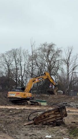 The scene shows a yellow Sany excavator working at a construction site on a gray, overcast day. The excavator is digging into a pile of dirt and mud, lifting a scoopful, and then dumping it. To the foreground's right, a rusty hydraulic breaker is lying on the muddy ground. Bare trees are visible in the background, along with some buildings and utility poles. A car drives by in the distance.