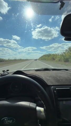 The video shows a first-person perspective from inside a Ford vehicle driving on a highway. The sun is high in the sky, casting bright light and glare through the windshield. The road ahead is straight with a clear lane in front. On the sides of the road, there are fields and trees. The interior of the car is visible, including the steering wheel with a Ford logo and the dashboard. There is a distant vehicle visible on the highway.