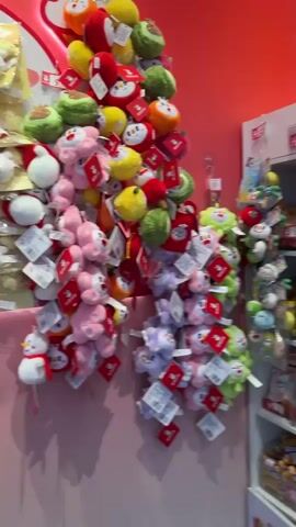 The video pans across a retail store, showcasing shelves stocked with a variety of merchandise. Primarily, the shelves are filled with packaged snacks and sweets, such as chips, crackers, and candy bars. Interspersed with the food items are decorative plush toys and other novelty items, many of which appear to be character-themed, featuring cartoon animals and possibly anime characters. Some of the plush toys are hanging from displays. The store's walls are painted in bright colors, including red and yellow, with cartoonish artwork visible on the upper sections. The floor is a speckled gray. A person is partially visible in the background, walking away from the camera towards the store's entrance, which is dimly lit and suggests an outdoor environment beyond. The overall impression is of a well-stocked novelty or convenience store, likely targeting a younger demographic or those interested in cute collectibles.