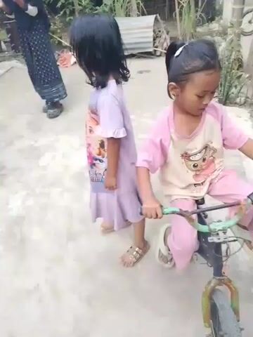 A short video shows a gathering of four people, two young girls and two women, in an outdoor setting. The scene appears to be a courtyard or yard, with a concrete floor and a backdrop of vegetation and simple structures, suggesting a rural or suburban environment.

In the foreground, a young girl in a pink shirt and pants is holding a yellow balloon. She is looking towards another young girl in a light purple dress. This second girl is partially turned away from the camera, and her face is not clearly visible.

In the background, a woman in a dark blue hijab and a patterned dress stands, appearing to observe the children. Another woman, mostly obscured, is also present.

The children seem to be engaged in play, with the girl in pink holding the balloon as if to offer it or play with it. The overall mood appears light and casual, with the woman in the hijab smiling, suggesting a friendly family gathering. The time of day appears to be daytime, with natural light illuminating the scene.

There is text visible on the purple dress of the second girl, but it is difficult to read clearly due to the video's resolution and the angle. It appears to be in a foreign script. The yellow balloon has some text and a cartoon face printed on it, but this is also not legible. The location context is provided as Meunasah Kulam, Indonesia.