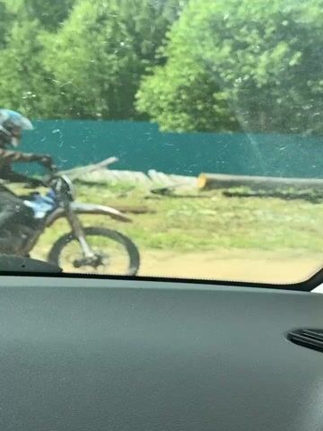 A person wearing a helmet and riding a dirt bike moves down a dirt path in a rural setting. The person is wearing a brown jacket and dark pants. The dirt bike is white and has knobby tires. The path is surrounded by green grass and trees. The sky is blue with some clouds. The video is shot from inside a car, looking out the side window. The car window frame is visible in the foreground. There is a wooden fence and some logs on the left side of the path. There are also power lines and utility poles visible in the background. The sun appears to be shining.