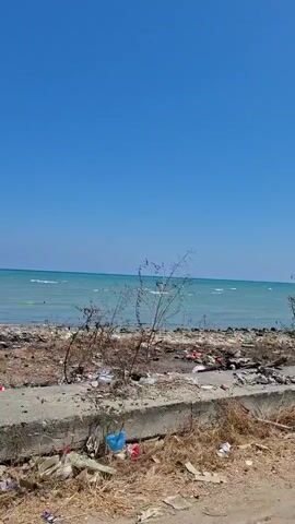 The video is a first-person perspective recording of a coastline. The sky is clear and blue. The ocean is a vibrant turquoise and appears to have moderate waves. The shoreline is littered with debris, including plastic bags, pieces of wood, and other refuse, indicating a significant amount of pollution. Concrete barriers and rubble are also visible along the coastline. In the distance, there are a few small trees and some structures that could be boats or buildings. A utility pole with wires is present in the foreground, indicating the presence of infrastructure. The recording pans slowly across the scene.