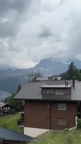 This media file captures a scenic view of a mountain village with a dramatic cloudy sky. The scene is set in Mürren, Switzerland, as indicated by the characteristic wooden chalets and the breathtaking mountainous backdrop. The primary subjects are the cluster of alpine-style houses with dark, sloped roofs, nestled on a grassy hillside. In the background, towering snow-capped mountains dominate the horizon, their rugged slopes partially obscured by dense green forests. A cable car system is visible, with its pylons and cables stretching across the valley, suggesting a mode of transport or a tourist attraction in the area.

The activity depicted is primarily observational; the viewer is presented with a static landscape. However, the cable car and a hint of red on the structure suggest potential movement or the operation of the cable car. The time of day appears to be daytime, likely afternoon, given the lighting. The weather is overcast, with heavy, grey clouds that create a somewhat dramatic and moody atmosphere. There are no discernible people in the foreground or mid-ground, but it's possible there are individuals on the cable car, though they are too distant to be identified.

Notable details include the architectural style of the houses, typical of Swiss mountain villages, and the imposing presence of the Alps. The contrast between the dark roofs of the houses and the green of the hills and dark trees, juxtaposed against the white snow and grey clouds, creates a visually striking image. The overall emotion evoked is one of awe at the natural landscape and perhaps a sense of peacefulness, despite the dramatic weather. There is no visible text in the media file.