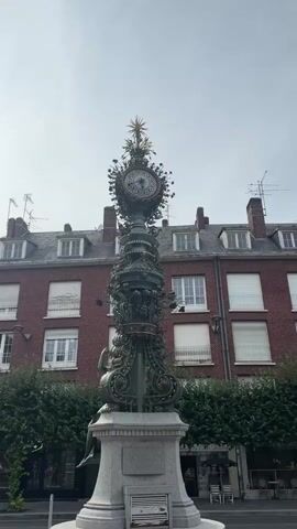 A vertical video captures a close-up shot of an ornate clock tower in an urban setting, then zooms out to reveal more of the surroundings. The clock tower is made of dark green metal with intricate decorative elements and a gold sunburst at its peak. Behind the tower, a red brick building with multiple windows and dormer roofs is visible. Lush green trees line the walkway in front of the building. A woman wearing a blue tank top and black pants walks across the foreground, carrying a backpack. The sky is overcast.