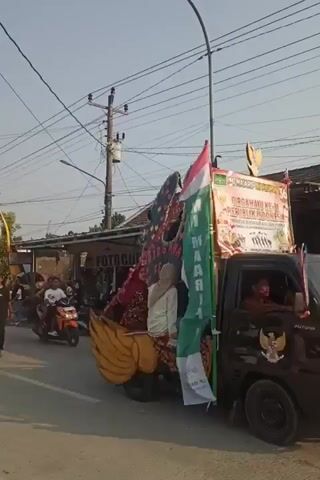 The video shows a street scene with a procession or parade. In the foreground, a young man in a black t-shirt and dark pants walks towards the camera. Behind him, a parade float shaped like a large bird with golden wings is moving down the street, carrying several people. The float is adorned with flags, including the Indonesian flag. To the left of the bird float, another float is visible, also decorated with banners and people. Further down the street, a truck with a banner that reads "PASAR DAHULAK" is part of the procession. The truck is decorated with gold accents and carries people. On the right side of the street, there are buildings, including one with a sign that says "FOTOC", and several motorcycles are parked. Power lines are visible overhead. People are gathered on the sidewalks watching the parade. The overall atmosphere appears festive.