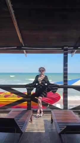 A woman stands on a wooden deck, leaning against a railing, with a beach and the ocean in the background. She is wearing a black cover-up over a bikini, sunglasses, and sandals. The deck has wooden lounge chairs. The ocean is calm with small waves. There are inflatable water toys on the beach. The sky is clear and blue.
