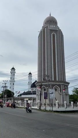The video captures a wide exterior shot of a street scene featuring a prominent mosque complex under an overcast sky. In the foreground, a distinctively tall, cylindrical minaret with a multi-tiered grey dome stands to the right of the paved road. This minaret is adorned with vertical architectural details in shades of grey and brown. Behind it, the main mosque building is visible, featuring a dark dome and two more traditional, slender white minarets with pointed tops.

The mosque complex is enclosed by a white, decorative fence, from which several Indonesian flags are flying. The street in front of the mosque is active with traffic. Several individuals are seen riding motorcycles (scooters); one person on a black scooter with a backpack is prominent in the mid-ground, moving from left to right. Other vehicles, including what appears to be a becak (cycle rickshaw), are also visible further down the road. Overhead, numerous utility poles and power lines crisscross the sky. Adjacent to the large minaret, there are informational signs or posters attached to a pole, but their specific text is not discernible.

The scene takes place during the daytime, with the uniformly grey sky indicating cloudy or overcast weather conditions. The presence of the mosque, the style of the minarets, the type of vehicles, and the Indonesian flags all point to a location in Indonesia, specifically Barat Yaman, as provided. The overall impression is that of a typical daily urban street scene.