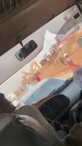The video shows a construction site viewed from inside a vehicle, possibly a bus or large van, with a driver visible in the foreground. The vehicle is in traffic, with other cars and people visible outside. The construction site includes heavy equipment, such as an excavator, and several workers, some wearing safety vests and hard hats. There is a pile of dirt or sand in the middle of the construction area. Smoke is visible coming from the excavator. The vehicle's dashboard and rearview mirror are visible, the rearview mirror appearing cracked. Passengers are visible in the vehicle. The lighting suggests it is daytime.