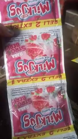 The video shows a close-up of a hand holding a chain of four small, pink, foil-wrapped packets of "Milk Jos" strawberry-flavored milk shake powder. The packets are stacked vertically, with the bottom two packets slightly out of focus. The packets have images of strawberries and a creamy drink. Text on the packets includes "Milk Jos," "Ice Blend," and "RASA STROBERI" (strawberry flavor). Some text in Indonesian is also visible, possibly indicating purchase or promotional information. The hand holding the packets is partially visible in the top left corner of the frame. The background is blurry but appears to be an indoor setting with wooden surfaces. The audio consists of someone speaking in Indonesian, expressing that the product tastes delicious.