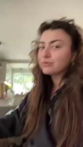 The video shows a woman with long, curly brown hair. She is wearing a dark gray hoodie and gold hoop earrings. Sunglasses are perched on her head. She says "Good morning." She then holds a brindle and white corgi up to the camera and tells the dog to say good morning. The corgi looks at the camera and licks its nose. The setting appears to be inside a home. The background shows a living room area with a couch, a window, and possibly a kitchen area in the distance.