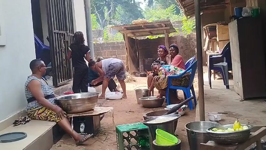 A group of women and one man are gathered in an outdoor space. One woman sits on the step of a building with a large metal bowl beside her. Two women are seated in chairs, working with metal bowls. A third woman and a man are bent over, working with plastic containers. There are several additional metal bowls scattered around, some with food items visible. A crude shelter stands in the background, along with a brick wall and trees. The area appears to be a courtyard or backyard, with a dirt ground. One of the women sitting on the chair is holding a baby. The woman on the step is wearing glasses and patterned clothing. The man is wearing shorts and a tank top.