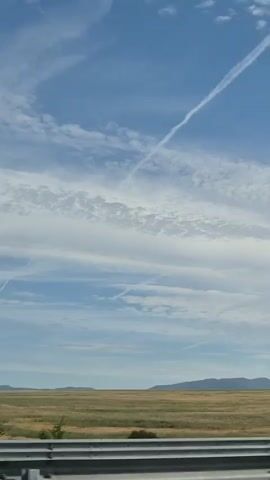 A vertical video pans up, showing a bright blue sky with wispy white clouds and a distinct contrail arcing across. Below the sky is a flat, golden-brown field, and in the foreground, a dark blue metallic surface with a silver stripe runs horizontally across the bottom of the frame. The scene appears to be captured from a moving vehicle, likely a truck or train, given the perspective and the metallic surface.