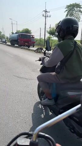 The video shows a person riding a scooter from behind, on a sunny day. The rider is wearing a helmet and a green and grey jacket. The scooter has a license plate that reads "L 3984 CAZ". The road is asphalt and there are trees and buildings in the background. The sky is blue and clear. The camera appears to be mounted on another scooter.