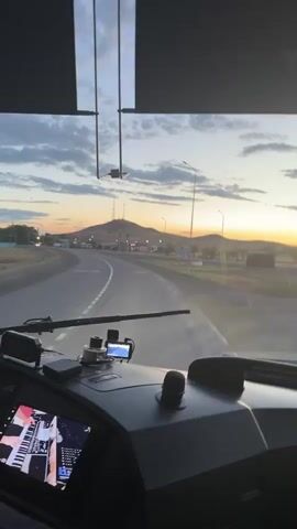 The video captures a view from inside a vehicle, likely a truck or bus, as it travels on a paved road. The perspective is from the driver's seat, looking out through the windshield. In the foreground, the dashboard is visible, featuring a large digital screen displaying what appears to be a music interface, possibly a virtual keyboard and sheet music. A GPS device is mounted near the windshield, and various buttons and controls are present on the dashboard.

The scene outside the vehicle depicts a rural or semi-rural landscape under a twilight sky. A winding road stretches ahead, bordered by fields and sparse vegetation. In the distance, a prominent hill or small mountain rises, topped with what looks like communication towers. Scattered lights and structures suggest a small settlement or roadside facilities along the road. The sky is dramatic, with a mix of dark clouds and streaks of orange and yellow light from the setting sun, indicating it is either dawn or dusk. The weather appears clear but overcast.

The activity taking place is vehicular travel, specifically a journey on a road. The time of day is clearly transitioning between day and night, given the colors of the sky. While no people are directly visible within the frame, the presence of a driver is implied by the viewpoint. The overall mood evoked is one of travel and contemplation, set against a visually striking natural backdrop. No legible text is discernible on the distant landscape, though there might be labels on the dashboard screens, which are too small to read clearly. The setting is Ulyanovskiy, Kazakhstan.