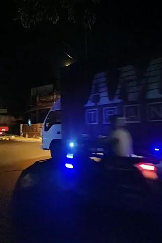 A nighttime video shows a large truck with the logo "AMS TRANS" on its side driving down a street. The truck has several blue lights under its chassis. The street is paved and illuminated by a street lamp and the truck's headlights. A building with a white facade and blue doors is visible on the right side of the street. A motorcycle is seen passing the truck. The truck's tires are visible, and the truck appears to be moving.