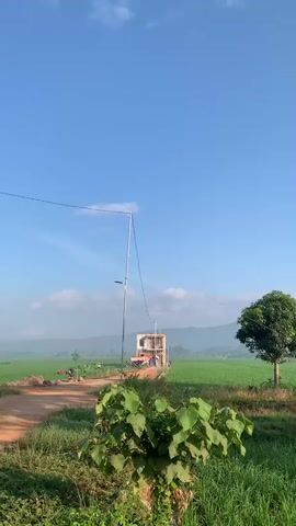 The video is a landscape shot of a rural area. In the foreground, there is green vegetation with some larger leaves. In the middle ground, there is a dirt path with some construction materials and a partially built structure with a tall antenna extending from it. A lone tree stands in a green field to the right of the construction. In the background, there is a vast green field, and beyond that, rolling hills under a clear blue sky with scattered white clouds. The scene appears to be peaceful and serene.