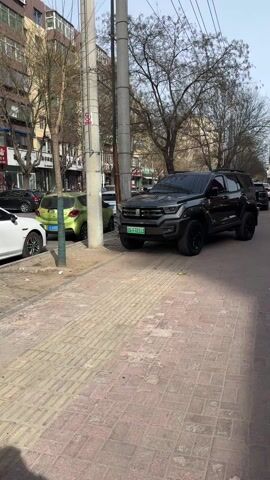 The video shows a dark gray SUV parked on a brick sidewalk next to a street in an urban environment.  The SUV has a distinctive front end design and large black tires. Other cars are parked nearby, and buildings and trees line the street.  The sky is visible in the background.