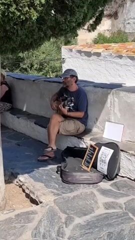 A man is sitting on a stone bench playing a guitar. He is wearing a blue t-shirt, khaki shorts, a baseball cap, and sandals. A guitar case is open on the ground in front of him, along with a small chalkboard sign and some papers. A woman is sitting on the bench to his left. The scene is outdoors with trees and a white wall in the background.