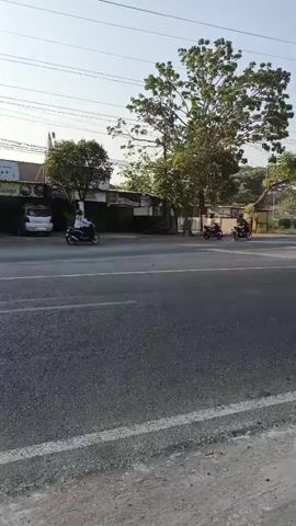 This is a video of a road with several motorcycles passing by. In the foreground, a man wearing a helmet and a dark shirt is riding a motorcycle with a child sitting behind him. The child is wearing a green and yellow jacket and has a backpack. They are riding on the right side of the road. In the background, other motorcycles are also visible on the road, some with riders wearing helmets. Trees and buildings line the side of the road, and the sky is clear and blue. The video appears to be taken from the side of the road, looking towards the traffic.