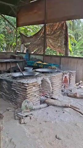 A short video displays an outdoor cooking setup. A large, circular metal pot rests on a brick structure that houses a fire. The pot appears to contain a liquid or food being stirred with a long utensil. Adjacent to the main cooking area, another metal pan sits atop a similar brick structure, also with embers visible. Two green plastic crates filled with what looks like yellow corn are positioned behind the cooking apparatus. The entire setup is under a makeshift roof covered with a patterned tarp. The ground is dirt and appears to have ash scattered around the brick structures. In the background, lush green foliage and trees are visible, suggesting a tropical or rural environment. A portion of a metal table or counter is seen to the left of the frame. A green, manual metal grinder is situated in front of the left side of the brick cooking structure.
