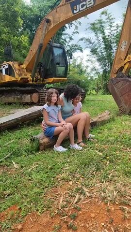 Three people, two young girls and a woman, are seated on logs in front of a yellow excavator. The woman is sitting between the two girls, with her arms around them. The girl on the left is wearing a t-shirt and denim shorts. The girl on the right is wearing a pink shirt. The woman is wearing a teal tank top and shorts. The excavator is large and yellow, with the JCB logo visible on its arm. The scene is outdoors, with grass and trees in the background. The lighting suggests it is daytime. The video is a short clip, showing the individuals posed for a photo.