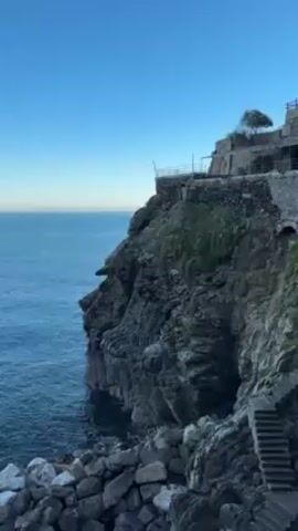 The video shows a rocky coastline meeting a calm blue sea under a clear, light-blue sky. A stone structure sits atop a cliff, with a small tree visible on the uppermost part. The camera pans from the cliff to the open water, then back to the rocky shoreline. Rocks and stairs lead down to the water's edge. There is no visible human activity in the video.