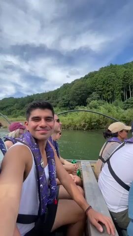 A group of people are on a boat traveling down a river. The man in the foreground is taking a selfie with a woman next to him. The boat has wooden benches and the people are wearing life vests. The river is surrounded by lush green trees on a hillside. The sky is cloudy with patches of blue.