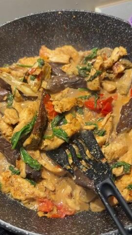 This close-up, overhead shot captures a culinary scene in a dark, non-stick frying pan. The pan is filled with a vibrant dish, seemingly in the process of being cooked. The main ingredients visible are thinly sliced pieces of chicken, chunks of dark purple eggplant, and leafy green vegetables, possibly spinach or basil. Interspersed among these are pieces of red chili or tomato, adding pops of color. The dish is coated in a creamy, orange-hued sauce that is bubbling, indicating it's on the heat.

A black spatula, with slotted tines, is partially submerged in the dish, suggesting active stirring or preparation. In the latter half of the clip, a stream of white liquid, likely coconut milk or cream, is being poured into the pan from above, creating a swirl of lighter color in the sauce. This indicates the dish is likely a curry or a similar saucy preparation, possibly Thai-inspired, given the common combination of ingredients and the use of coconut milk.

The lighting is bright and focused on the pan, suggesting it's being filmed in a well-lit kitchen environment. There is no visible text in the media. The overall impression is one of delicious food being prepared, with a focus on the textures and colors of the ingredients and the creamy sauce. The lack of any background context or people makes it difficult to pinpoint a specific location beyond a kitchen setting. The provided location context "Kokkorevo, Russia" does not offer specific visual cues within the frame itself.