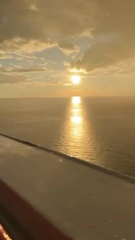 This is a vertical, low-angle video of a body of water during what appears to be a sunset or sunrise. The sun is visible as a bright orb low on the horizon, casting a shimmering golden reflection across the water's surface. The sky is filled with clouds, some of which are illuminated by the sun's glow, while others are darker and more defined. In the foreground, there is a railing or edge of a structure that appears to be wet, with small droplets of water visible on its surface. The overall mood of the video is serene and tranquil.
