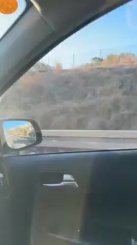 The video is taken from inside a car driving on a highway. The sun is shining brightly, almost directly ahead, partially obscured by a hill with vegetation. Electrical transmission towers are visible in the distance. The dashboard of the car is visible, with a sticker on the passenger side. A reflection of the road behind is visible in the side mirror. The driver's hands are occasionally visible on the steering wheel. The car's infotainment system is on.