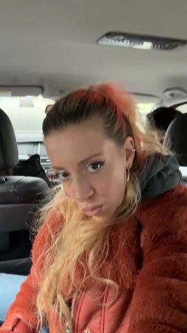 The video shows a blonde woman in a car, wearing an orange fuzzy jacket and silver hoop earrings. She has red streaks in her hair, which is styled in a high ponytail. She is wearing eye makeup and lipstick. She makes several facial expressions, including pursed lips, a wink, and sticking her tongue out. She also makes a rock-and-roll hand gesture. The interior of the car is visible, including the dashboard, rearview mirror, and other seats. Music plays in the background.