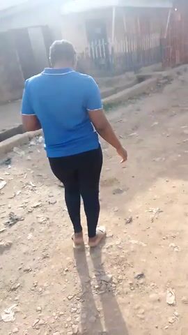 A woman walks on a dirt road littered with trash. She has dark hair pulled back, wears a blue t-shirt with white accents, black leggings, and sandals. She looks at her phone as she walks. A concrete drainage ditch runs parallel to her path. Buildings are visible in the background.