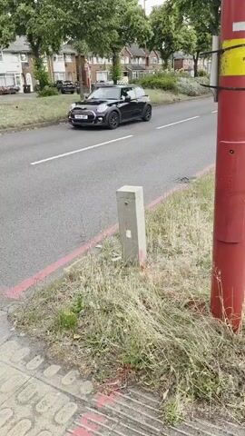The video shows a roadside scene. In the foreground, there is a red pole with a yellow top, a concrete bollard, and a textured sidewalk. Beyond the sidewalk, there is a grassy verge separating the sidewalk from a road. The road has a red line painted near the edge. Cars are visible driving on the road. Further down the road, houses and trees line the street. The pavement has yellow and red markings, possibly indicating measurements or construction areas. A dark-colored car is parked on the side of the road in the distance, and a blue SUV is driving on the road.