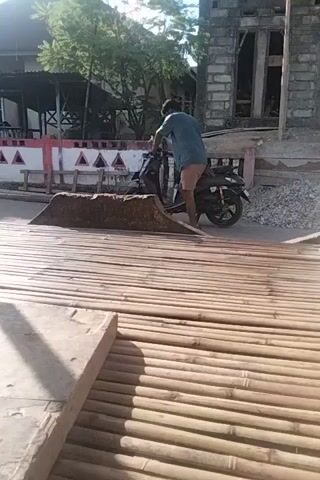 The video pans from right to left across an outdoor area. The ground appears to be a rough concrete surface with some scattered shadows. To the left, a building structure with a tin roof is visible. There are wooden beams supporting the roof and what looks like a wooden plank walkway. In the background of the building structure, there is a person wearing a red shirt. Some laundry is hanging on a clothesline to the left of the walkway. White fishing nets are piled up on the right side of the image. The lighting suggests it is daytime, with strong sunlight creating distinct shadows.