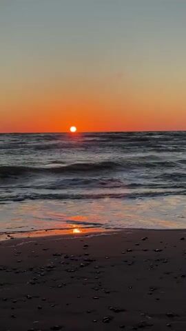 A vertical video shows a sunset over the ocean. The sun, a bright orange orb, is low on the horizon, casting a warm glow across the sky and water. Waves crash gently onto a sandy beach in the foreground, their white foam reflecting the sunset's colors. The beach is littered with small pebbles and shells. The overall mood is peaceful and serene.