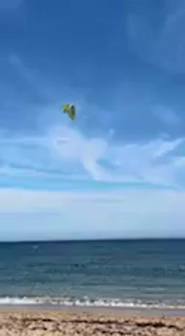 A bright yellow and green kitesurfing kite soars high in a clear blue sky, marked by wispy white clouds. Below, the deep blue waters of the ocean stretch out, meeting a sandy beach in the foreground. A kitesurfer is visible in the water, positioned to ride on their board. The kite drifts gently across the frame, propelled by the wind, while the kitesurfer navigates the calm waves. The scene captures a sunny day, likely ideal for watersports, in Barbate, Spain.