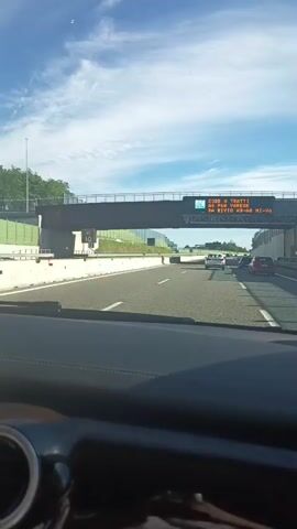 The video, filmed from the perspective of a driver, shows a daytime scene on an Italian highway near Luisago. The initial shot captures the view through the windshield, revealing multiple cars on the road ahead and an overhead electronic sign spanning the motorway. The sky is bright with some clouds, indicating clear weather.

The prominent electronic sign displays text in white capital letters on a dark background. It reads:
"CODE A TRATTI" (Queues in sections)
"SS. 336 VARESE" (State Road 336 Varese)
"DA BIVIO SS-36 MI-VA" (From Junction State Road 36 Milan-Varese)
This indicates traffic information and navigational details for the area, specifically mentioning potential queues and directions towards Varese and the junction of State Road 36 leading to Milan-Varese.

The camera then pans slightly to the right, showing the interior of the car's A-pillar and the bright sunlight reflecting off the window and interior surfaces. The view outside still shows the highway and the surrounding environment, including a concrete barrier and some trees. The video primarily depicts a car journey on a sunny day.