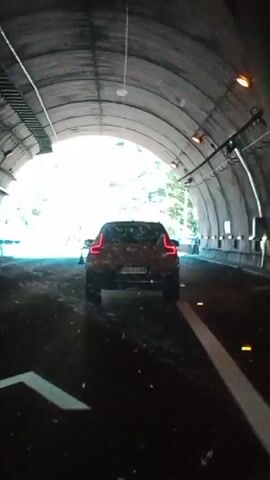 The video begins inside a dark tunnel, showing the rear of a white SUV driving away from the camera. As the SUV moves towards the tunnel exit, the light from outside begins to illuminate the scene. Beyond the tunnel, a mountainous landscape with sparse vegetation is visible. Other vehicles, including a truck and several cars, are present on the highway. The SUV exits the tunnel and continues on the highway.