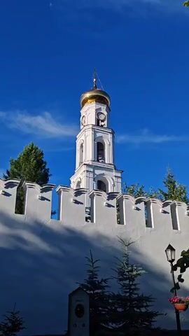 A vertical video shows a bell tower with a golden dome and a cross atop it. The tower is white with arched windows and a clock face. It is partially obscured by a white wall with battlements, behind which are trees. The sky is a clear, deep blue with a few wispy clouds. There is a street lamp on the right side of the frame, with a potted plant beneath it. The overall impression is of a sunny day.