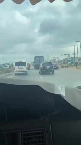 The video captures a view from inside a vehicle moving along a multi-lane road. The sky is overcast. In front, there is a truck carrying a large blue shipping container, followed by a black SUV, and then a black hatchback. To the right, a yellow minibus is visible, as well as a three-wheeled vehicle commonly known as an auto-rickshaw. A person is seen walking on the sidewalk next to a row of flags. The road appears to be in a commercially developed area with buildings and infrastructure lining the sides. The vehicle the footage is being filmed from has a textured dashboard and a strip of tape.