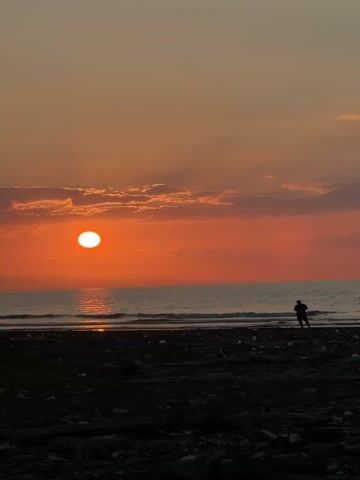A silhouette of a person stands on a dark, debris-strewn beach facing the ocean during a vibrant sunset. The sun, a bright orange orb, is positioned low on the horizon, casting a warm glow across the sky and reflecting on the water. Wispy clouds, tinged with pink and orange, drift across the upper half of the frame. The ocean waves gently lap at the shore. The overall scene depicts a serene and colorful end to the day.