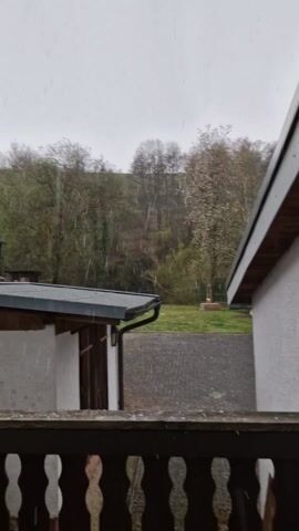 The video shows a view from a balcony, during what appears to be a hailstorm or a mix of rain and snow. The balcony has a wooden railing with decorative cut-outs, and a wooden floor. The pellets or snowflakes are visible as they fall and accumulate on the wooden planks. Beyond the balcony, there is a view of a residential area with buildings, a paved area, and a grassy yard. In the background, there are trees on a slope. The sky is overcast and gray.