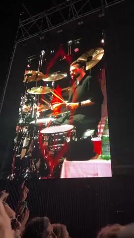 A bald man with a muscular build, wearing a dark t-shirt and dark pants, plays a bass guitar on a large screen. The screen displays abstract, colorful graphics behind him, shifting between red, purple, and blue hues. The lighting on the screen is dramatic, with spotlights highlighting the musician. Below the screen, in the lower part of the frame, a crowd of people can be seen, with their hands raised in the air, indicating they are at a concert. The overall environment appears to be a dark concert venue.