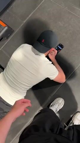The video begins with a close-up of a person lying on a dark gray floor, wearing a white t-shirt, black shorts, and a black baseball cap. They are holding a smartphone in their hands, with the screen displaying a timer. The camera angle shifts to show another person's legs and feet, wearing black pants and white sneakers. The scene appears to be in a gym, indicated by the presence of gym equipment like weight racks and benches in the background. The lighting is bright, and the environment looks like an indoor fitness facility. The video is short, with the scene changing rapidly.